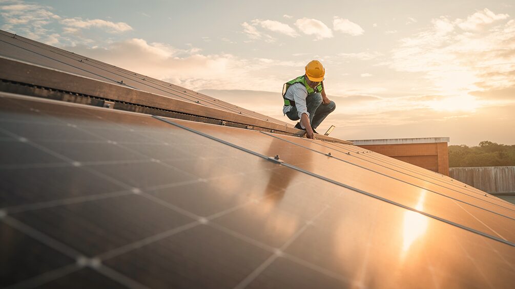 Person mit Helm befindet sich bei Sonnenuntergang auf einem Dach und bringt Solaranlagen an